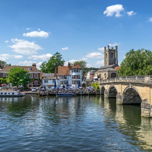 Knight Frank Henley-on-Thames Estate Agents - An historic stone bridge, leading toward a traditional waterfront town with a prominent church tower under a blue sky with scattered clouds.