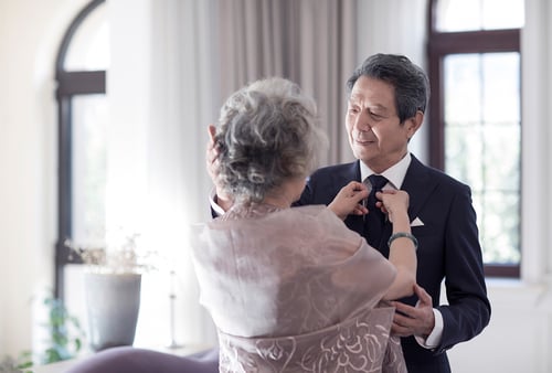 Elderly couple in a softly lit room; woman adjusts man's tie affectionately. Both are elegantly dressed, conveying warmth and love.