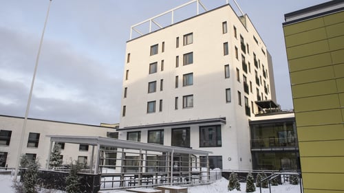 Ulkokuva Myllystä. Kerrostalorakennus on vaalea, ja sen edessä on katettu ulkotila, jossa on penkkejä. Pihapiirissä on pieniä havupuita.