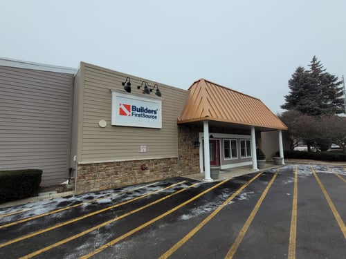 A photo of the Builders FirstSource storefront in Auburn Hills, MI, during winter. The building features light beige siding with a stone facade along the lower portion and a copper-colored metal awning supported by white pillars above the entrance. The company’s logo is prominently displayed on a white sign with black overhead lighting fixtures. The parking lot in the foreground shows snow lightly scattered across the asphalt, with yellow parking lines visible. Leafless trees and evergreen shrubs frame the building under an overcast sky.