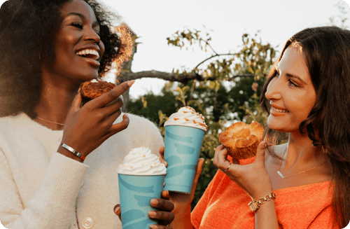 Two women outdoors, smiling at each other while holding pastries and Caribou cups.