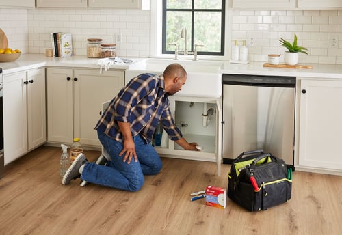Man placing a water leak/freeze detector under a kitchen sink. The product box and tools are on the floor nearby.