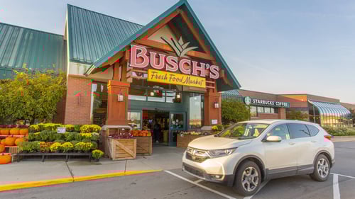 White SUV driving past Busch's Fresh Food Market with plants and pumpkins sitting outside