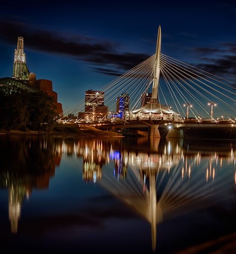 A serene evening view of a cable-stayed bridge with bright city lights creating shimmering reflections on the calm river, against a deep blue sky.