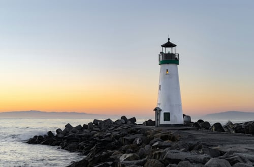 Lighthouse at sunrise with a soft gradient sky, standing on a rocky shore. The calm sea reflects hues of orange and blue, conveying tranquility.