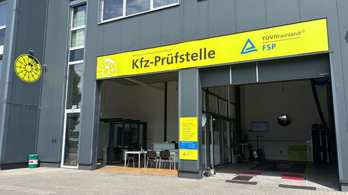 Das Bild zeigt die Außenansicht einer Kfz-Prüfstelle mit großem gelbem Schild, auf dem "Kfz-Prüfstelle" sowie die Logos von TÜV Rheinland und FSP stehen. Links ist eine gelbe Prüfplakette an der Fassade, der Eingangsbereich ist offen und es stehen Tische und Stühle davor.