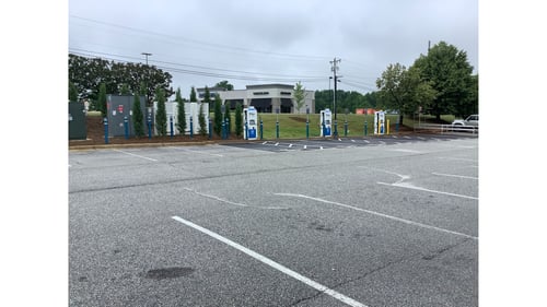 Several EVgo fast charging units installed in marked parking spaces at a public charging station, with EVgo logos on the chargers, protective bollards around each unit. NACS and CCS chargers featured in the image.