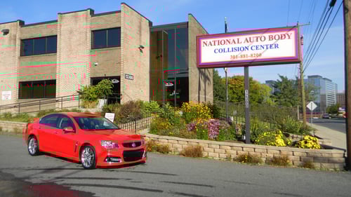 The image shows the exterior of “National Auto Body Collision Center,” a two-story brick building with large windows. A bright red car is parked in front of the building near a landscaped area with colorful flowers. A large sign with the business name and phone number is prominently displayed on a pole next to the building, facing the street. The sign reads “NATIONAL AUTO BODY COLLISION CENTER” in red and blue letters, with the phone number in red below. The scene is bright and sunny, highlighting the clean and professional appearance of the facility.