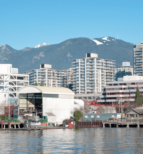 Waterside urban landscape with modern high-rise buildings against a backdrop of snow-topped mountains, under a clear blue sky. Boats in the foreground.