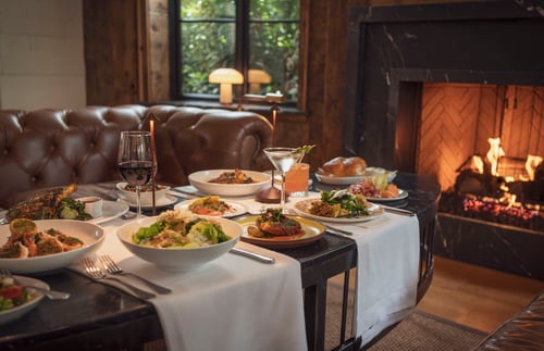 Elegant dining spread at The Farm Italy featuring Italian dishes, cocktails, and wine beside a glowing fireplace, creating a warm and inviting atmosphere perfect for date nights and special occasions.