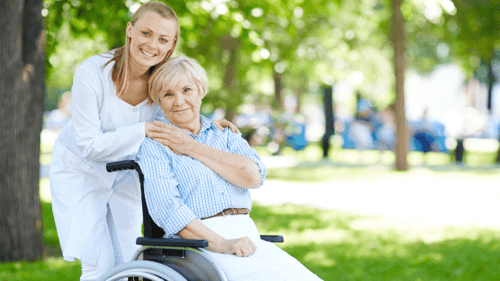 A smiling caregiver in a white coat stands beside a senior woman in a wheelchair outdoors, resting a hand on her shoulder in a sunlit park.