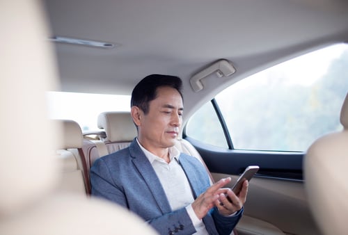 A man in a blue blazer sits in the backseat of a car, focused on his smartphone. The mood is calm and professional, with soft daylight filtering in.