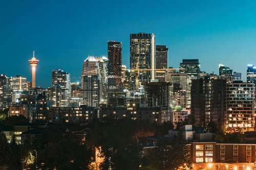 Calgary downtown skyline at dusk featuring the illuminated Calgary Tower and modern skyscrapers including the distinctive Bow Tower, with city lights glowing against a twilight blue sky.