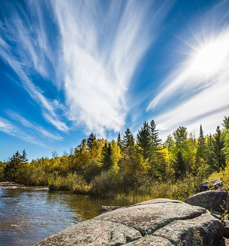 Sunlit forest with vivid autumn foliage by a rocky riverbank. Dramatic wispy clouds stretch across a clear blue sky, conveying a serene atmosphere.