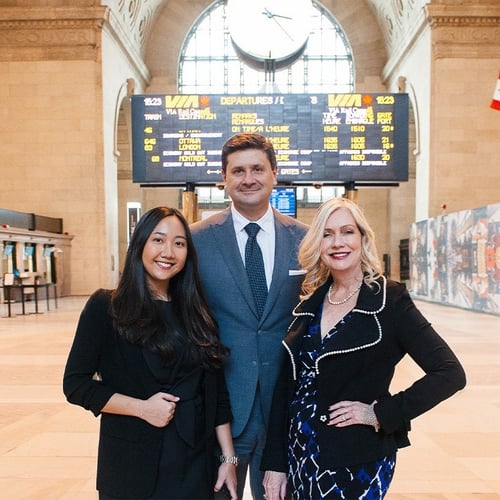 Three business professionals standing in Union station hall with a large departures board visible above them. The woman on the left wears a black blazer, the man in the center wears a blue suit and tie, and the woman on the right wears a black blazer and a patterned blue dress. The ornate station features a circular clock, wooden ceiling details, ticket windows on the left, decorative wall murals on the right, and Canadian flags displayed on the upper right wall.