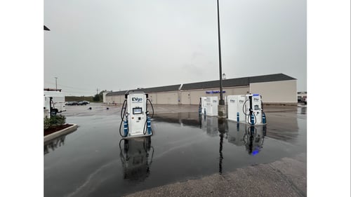 Several EVgo fast charging units installed in marked parking spaces at a public charging station, with EVgo logos on the chargers, protective bollards around each unit.