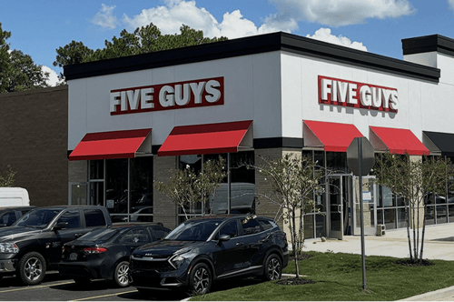 Exterior of a Five Guys restaurant with red awnings and parked cars in front.