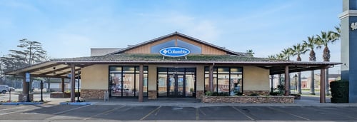 Photograph of Columbia Sportswear storefront with blue and white logo sign above the entrance. The large glass windows display mannequins dressed in outdoor clothing, and the store's interior is visible.