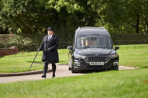 A Coop funeral director wearing a black top hat, frock coat and cane walks ahead of a black hearse towards the funeral location.