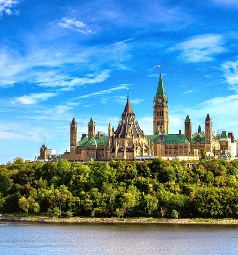 Majestic Gothic Revival architecture of Ottawa's Parliament Hill towers above lush green trees and river under a clear, vibrant blue sky.