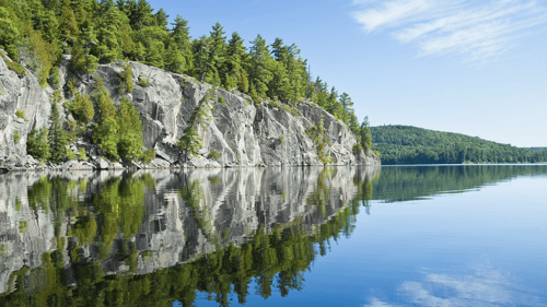 Scenic view of rocky cliffs adorned with lush green trees, reflected on the tranquil, mirror-like surface of a serene blue lake under a clear sky.