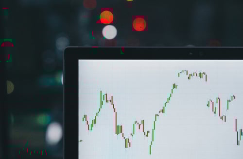 Close-up of a computer screen displaying a fluctuating stock market graph with green and red lines. Blurred background with bokeh lights, dark ambiance.