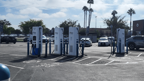 Several EVgo fast charging units installed in marked parking spaces at a public charging station, with EVgo logos on the chargers, protective bollards around each unit.
