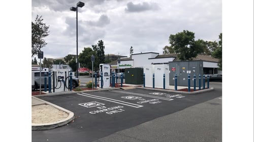 Several EVgo fast charging units installed in marked parking spaces at a public charging station, with EVgo logos on the chargers, protective bollards around each unit.