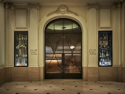 A dimly lit lounge bar with rich red velvet seating, dark wooden tables, and candlelit ambiance. The back wall displays a wide variety of bottles, creating a cozy, intimate setting.