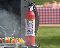 A small fire extinguisher sits on a table extension of a grill, there is smoke rising and a plate of kebabs next to it.