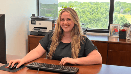 Woman with long blonde hair sitting at an office desk using a keyboard and mouse, window with blinds and green trees outside in background.