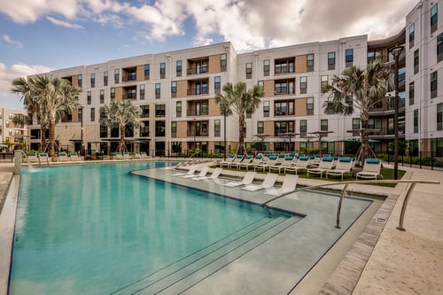 Outdoor swimming pool with in-water lounge chairs, surrounding chaise lounges, and palm trees.