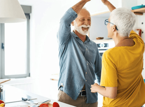 An elderly couple smiling and dancing together in their kitchen.