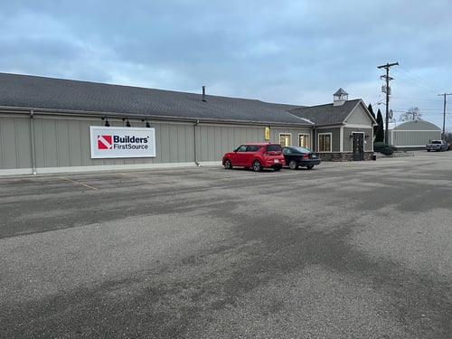 The front view of a Builders FirstSource lumber yard with a clean parking area and the company logo prominently displayed on the building’s exterior. A few cars are parked in front, and the structure features a cupola-style architectural element, with overcast skies overhead.