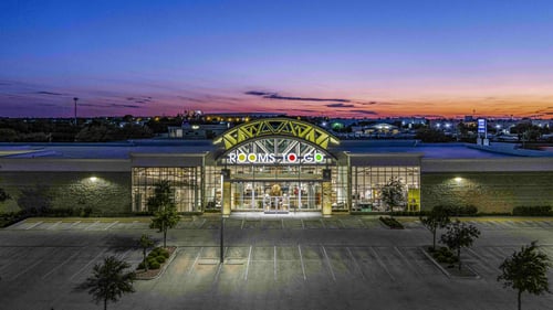 The exterior view of Rooms To Go Round Rock, TX, captured at sunset with the store’s illuminated sign and glass entrance. The parking lot is empty, and the colorful sky creates a picturesque backdrop for the store.