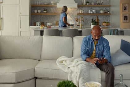 Man sitting on a couch looking at the Kidde App while woman is cooking in the background