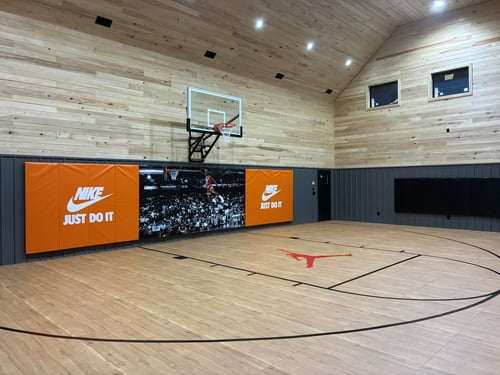 A professional indoor basketball court with light wood walls and flooring, featuring orange wall pads and a red Michael Jordan silhouette.