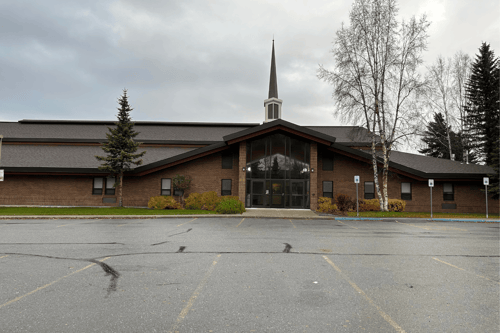 Front view of the Baxter church building showing the south entrance with large glass doors and tall windows beneath a peaked roofline. A steeple rises above the center, with trees, landscaping, and parking spaces visible in the foreground under an overcast sky.