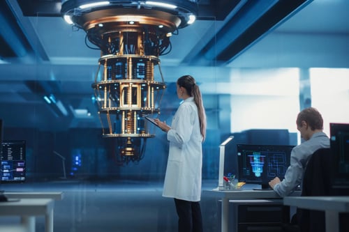 Two scientists in a high-tech laboratory observe a glowing cylindrical quantum computer suspended from the ceiling while monitoring data on screens.