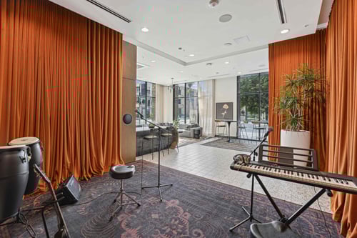 Music performance space with orange drapes, a microphone, keyboard, congas, and a stool on a patterned carpet, opening to a bright lounge.