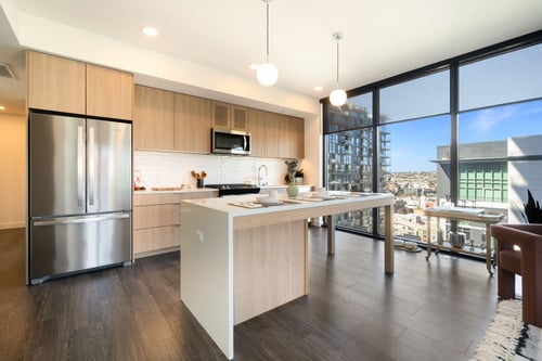 Modern kitchen with light wood-style cabinets, white island, stainless steel appliances, and city views through large windows.