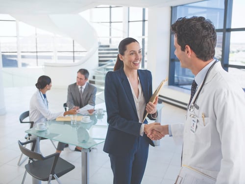 A doctor shakes hands with a smiling businesswoman holding a file in a bright office. In the background, two people converse at a glass table.