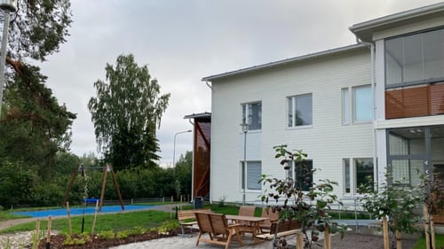 Ulkokuva Säteen pihapiiristä. Rakennus on valkoinen. Pihalla on kivitetty alue, jolla on pöytäryhmä. Alueella on lisäksi keinu, nurmikkoa ja istutuksia.
