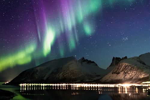 Aurora borealis with green and purple lights above mountains and illuminated dock reflections.