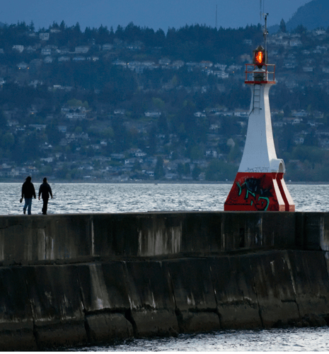 Two people walk along a concrete pier towards a small lighthouse, set against a backdrop of water and distant forested hills under a gray sky.