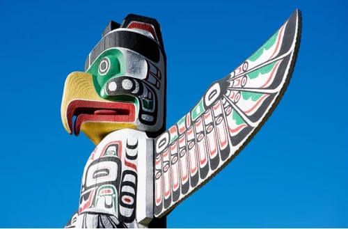 Mât totémique coloré mettant en vedette un oiseau stylisé avec des motifs noirs, rouges et verts complexes contre un ciel bleu clair, transmettant la richesse culturelle.