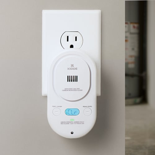 A carbon monoxide alarm plugged into the wall with the display illuminated in blue that shows "CLR" for clear.