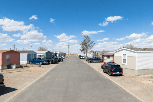 A residential street with houses on both sides and cars parked on the right side at Queens MHC, in El Paso, TX