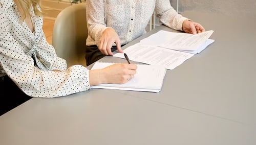Two people review documents at a gray table. One person points, and the other writes. Both wear polka dot shirts, conveying a focused atmosphere.