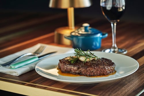 A perfectly cooked steak topped with garlic and rosemary on a white plate, served alongside a glass of red wine and a small blue pot on a wooden table.